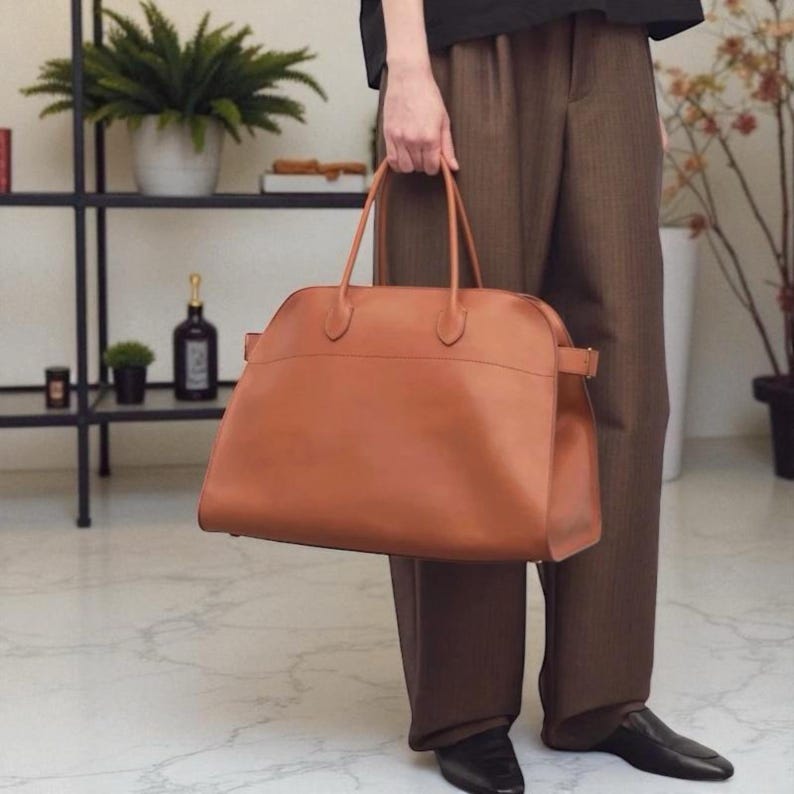 May include: A tan leather handbag with two top handles and a rectangular shape. The bag has a buckle closure on the side. The bag is a light brown colour and is being held by a person.