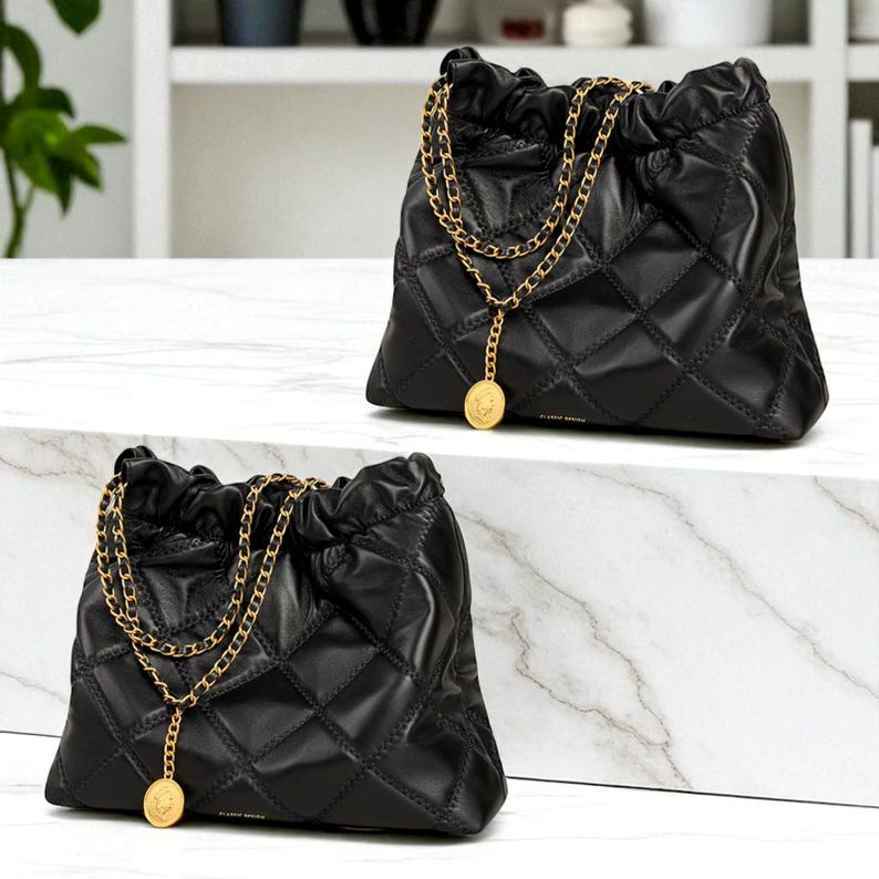 Puede incluir: Dos bolsos de mano de cuero negro acolchado con correas de cadena doradas y colgantes de monedas. Los bolsos tienen un cierre de cord&oacute;n y un patr&oacute;n de diamantes. Los bolsos se exhiben sobre una superficie de m&aacute;rmol blanco.