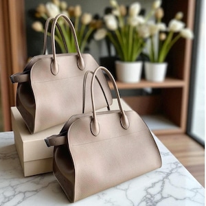 May include: Two taupe-coloured handbags with rounded shapes and top handles are displayed. The bags have a structured design with a textured surface. They are placed on a marble-patterned surface, with a neutral-toned box beneath one.