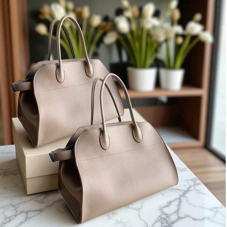May include: Two taupe-coloured handbags with rounded shapes and top handles are displayed. The bags have a structured design with a textured surface. They are placed on a marble-patterned surface, with a neutral-toned box beneath one.