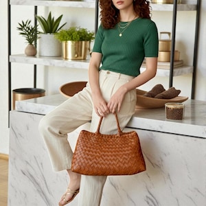 May include: A woman wearing a green short-sleeved top and cream-colored pants, holding a large, brown woven leather tote bag. The bag has two handles and a rectangular shape. The background includes shelves with plants and decorative items.