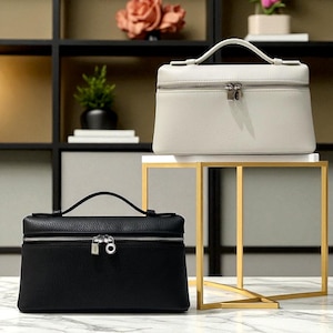Puede incluir: Dos neceseres de cuero, uno blanco y otro negro, con cremalleras y asas plateadas. Las bolsas son rectangulares con un asa superior y se exhiben sobre un soporte dorado y blanco. Las bolsas están sobre una superficie de mármol.