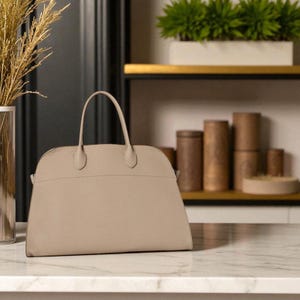 May include: A beige leather handbag with a structured design. The bag has a curved top, a top handle, and a zip closure. The bag is on a marble countertop.