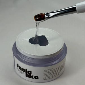 Puede incluir: Un frasco de gel transparente con un pincel, probablemente para uñas. El gel gotea del pincel. El frasco tiene una tapa blanca con un agujero negro y las palabras "feels like" impresas en el lateral.