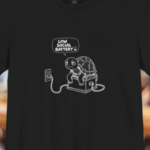 Peut inclure: T-shirt noir avec un dessin blanc d'une tortue branchée sur une prise électrique. Le texte "LOW SOCIAL BATTERY" est affiché dans une bulle de dialogue, au-dessus de la tortue.