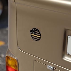 May include: A circular sticker featuring a distressed American flag design with a black and gray color scheme. The flag includes a yellow stripe and a black border, affixed to a tan vehicle surface.