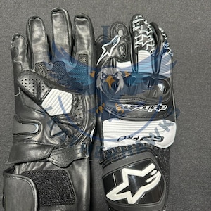 Könnte beinhalten: Paar schwarze und weiße Motorradhandschuhe. Die Handschuhe bestehen aus Leder und synthetischen Materialien mit Schutzpolstern an Knöcheln und Handgelenk. Die Handschuhe tragen die Marke "Alpinestars" und "GP Pro".