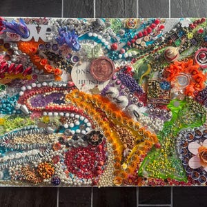 Pode incluir: Uma obra de arte em técnica mista com fundo branco, apresentando uma colagem de contas, botões e outros pequenos objetos coloridos. A palavra "LOVE" está escrita em letras laranja. A obra é uma exibição vibrante de texturas e cores.