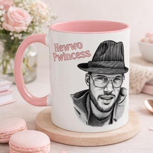May include: A white ceramic mug with a pink handle and interior rim. The mug features a black and white sketch of a person wearing a pinstriped fedora and glasses, with the red text "Hewwo Pwincess" printed above the illustration.
