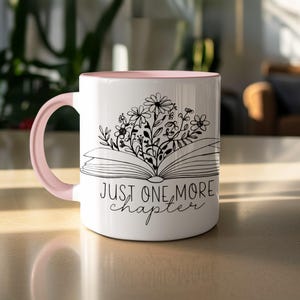 May include: White ceramic mug with a pink interior and handle. The mug features a black and white illustration of flowers and an open book. The text "JUST ONE MORE chapter" is written below the book.