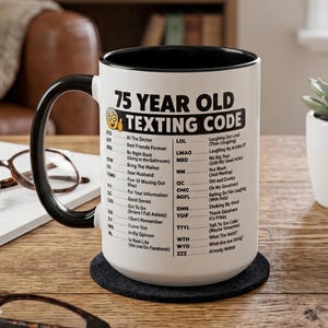 May include: A white ceramic mug with a black interior and handle, featuring the black printed text 75 YEAR OLD TEXTING CODE and a list of humorous acronym definitions including ATD for At The Doctor, BFF for Best Friends Forever, and ZZZ for Already Asleep.
