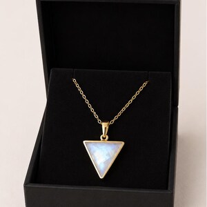 Op de afbeelding: Een goudkleurige ketting met een driehoekige hanger, gepresenteerd in een zwarte sieradendoos. De hanger heeft een lichtblauwe steen in een gouden frame, hangend aan een fijne ketting.