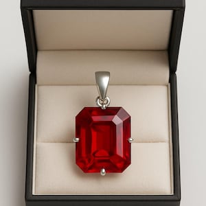 Op de afbeelding: Een rode, smaragdgeslepen hanger met een zilveren balein, gepresenteerd in een open zwarte sieradendoos met een crèmekleurig interieur. De hanger is een levendige, gefacetteerde edelsteen, die zijn rechthoekige vorm en reflecterende eigenschappen laat zien.
