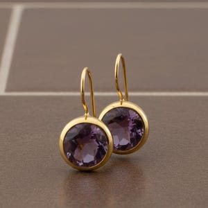 May include: A pair of gold-colored earrings with large, round, purple gemstones. The earrings have a simple, elegant design, with the gemstones set in a gold bezel. The earrings are displayed on a brown surface.