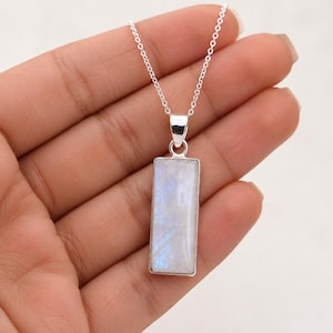 Peut inclure: Un pendentif rectangulaire en pierre de lune serti dans une monture en argent, suspendu à une fine chaîne en argent. La pierre de lune présente un aspect blanc laiteux translucide avec de subtils reflets bleutés irisés sur sa surface plate.
