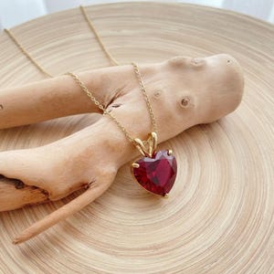 Puede incluir: Un collar dorado con un colgante de gema roja oscura en forma de corazón. El colgante está facetado, captando la luz. El collar se exhibe sobre un trozo de madera clara, sobre un fondo de madera.