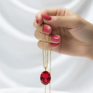 May include: A gold chain necklace featuring a large, oval, red gemstone pendant. The necklace is held by a hand with red nail polish against a white, draped backdrop. The pendant is the central element.