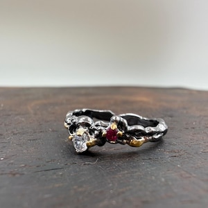 May include: A silver ring with a textured design, featuring a clear gemstone and a small red gemstone. Gold accents are visible around the gemstones. The ring is set against a dark wooden surface.