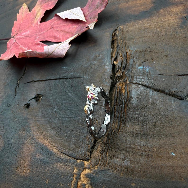 May include: A silver ring with a floral design, featuring small gold accents and a red gemstone. The ring is set against a dark wooden surface, with a red leaf in the background. The ring's design is intricate and detailed.