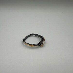 May include: A black and gold ring with a textured band. The ring features a small red gemstone and a clear gemstone. The ring is set against a white background.