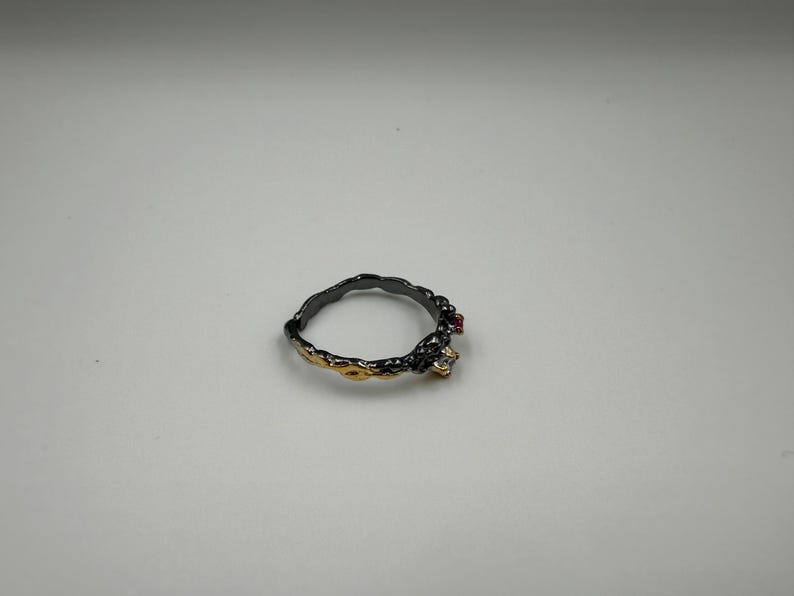 May include: A black and gold ring with a textured band. The ring features a small red gemstone and a clear gemstone. The ring is set against a white background.