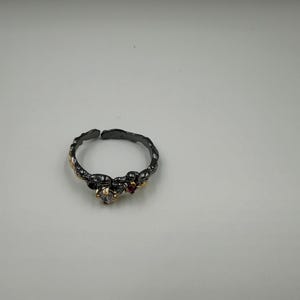 May include: A dark gray ring with a textured band, featuring small gold accents and clear and red gemstones. The ring has an open design, allowing for slight size adjustments. A piece of jewelry.