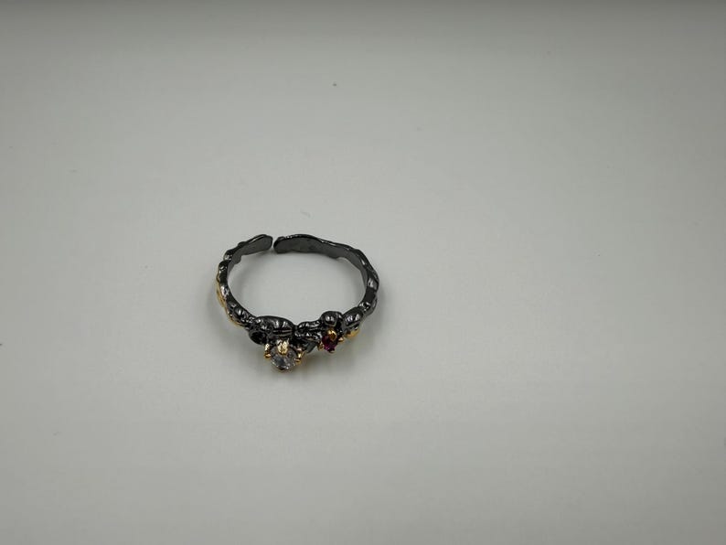 May include: A dark gray ring with a textured band, featuring small gold accents and clear and red gemstones. The ring has an open design, allowing for slight size adjustments. A piece of jewelry.