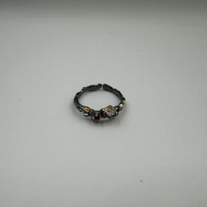 May include: A dark silver adjustable ring with gold accents and three gemstones. The ring features a small, round, clear gemstone, a small red gemstone, and a gold accent. The ring is set against a white background.