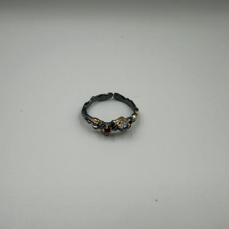 May include: A dark silver adjustable ring with gold accents and three gemstones. The ring features a small, round, clear gemstone, a small red gemstone, and a gold accent. The ring is set against a white background.