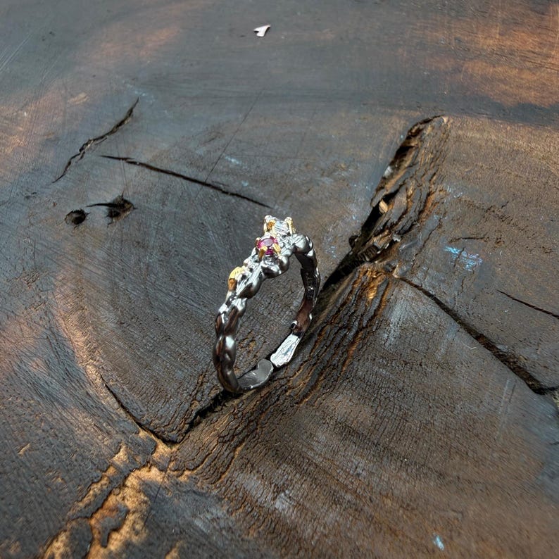 May include: A silver ring with a textured band and a cluster of small, pink gemstones. The ring has gold accents and sits on a dark wooden surface, showcasing its intricate design and craftsmanship. The ring's design is inspired by nature.