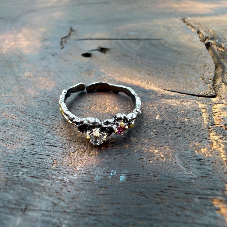May include: A silver-toned ring with a textured band and two small gemstone clusters. One cluster features a clear stone, the other a small red stone. The ring is set on a rustic wooden surface.