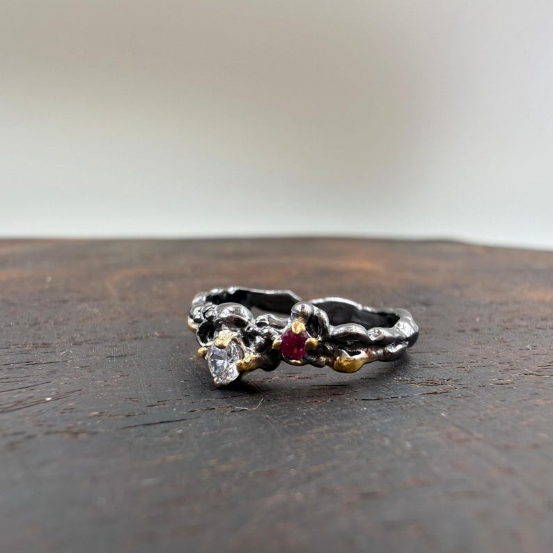 May include: A silver ring with a textured design, featuring a clear gemstone and a small red gemstone. Gold accents are visible around the gemstones. The ring is set against a dark wooden surface.