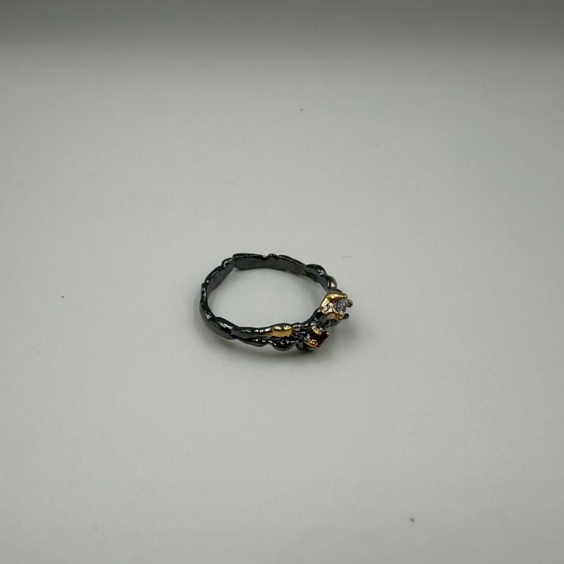 May include: A dark gray ring with a textured band. The ring features a gold accent and is adorned with a small, clear gemstone and a red gemstone. The ring is set against a white background.
