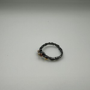 May include: A dark gray ring with a textured band and small gemstones. The ring has a unique, organic design, with a rough, almost bark-like texture. The gemstones are a mix of clear, red, and yellow.