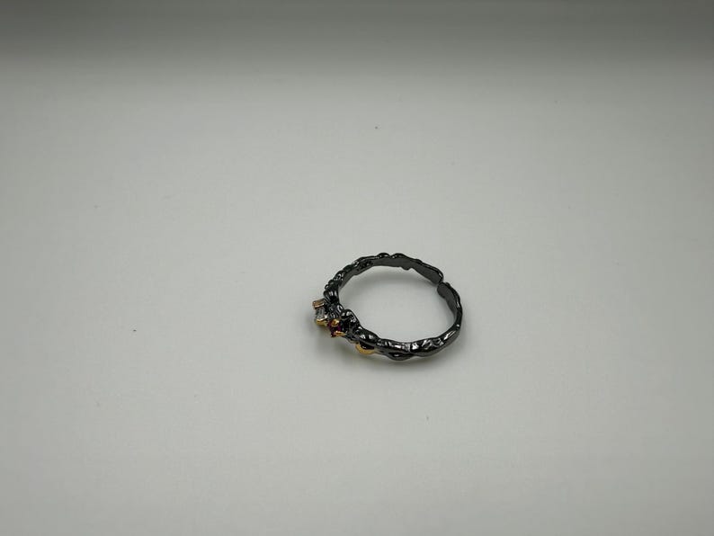 May include: A dark gray ring with a textured band and small gemstones. The ring has a unique, organic design, with a rough, almost bark-like texture. The gemstones are a mix of clear, red, and yellow.