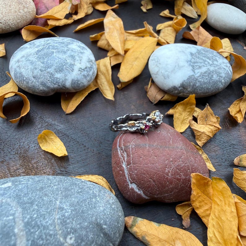 May include: A silver ring with gold accents and small red and clear gemstones. The ring is displayed on a red rock, surrounded by gray stones and dried yellow leaves. The ring's design is intricate and detailed, showcasing a unique jewelry piece.