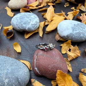 May include: A silver ring with gold accents, featuring a clear gemstone and two red gemstones. The ring is displayed on a red stone, surrounded by gray stones and yellow leaves, creating a natural, earthy aesthetic.