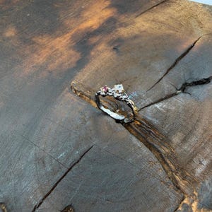 May include: A silver ring with a unique, textured design. The ring features a cluster of small gemstones, including a clear, round stone and a smaller pink stone. The ring is set against a rustic, wooden background, highlighting its handcrafted appearance.