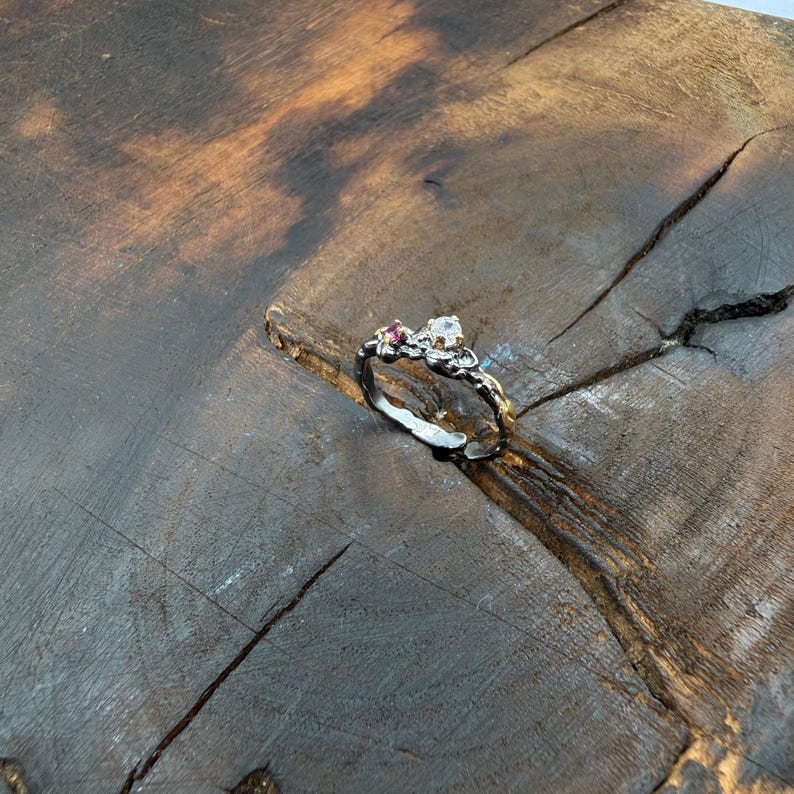 May include: A silver ring with a unique, textured design. The ring features a cluster of small gemstones, including a clear, round stone and a smaller pink stone. The ring is set against a rustic, wooden background, highlighting its handcrafted appearance.