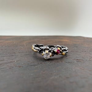 May include: A silver ring with a textured band, featuring a clear round gemstone and a small red gemstone. The ring has gold accents and a unique, organic design, perfect for a special occasion.