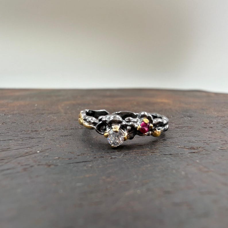 May include: A silver ring with a textured band, featuring a clear round gemstone and a small red gemstone. The ring has gold accents and a unique, organic design, perfect for a special occasion.