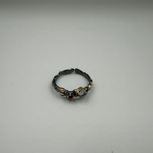 May include: An adjustable ring with a dark, textured band. The ring features gold accents and three gemstones: a clear, round stone and a small red stone. The ring is set against a white background.