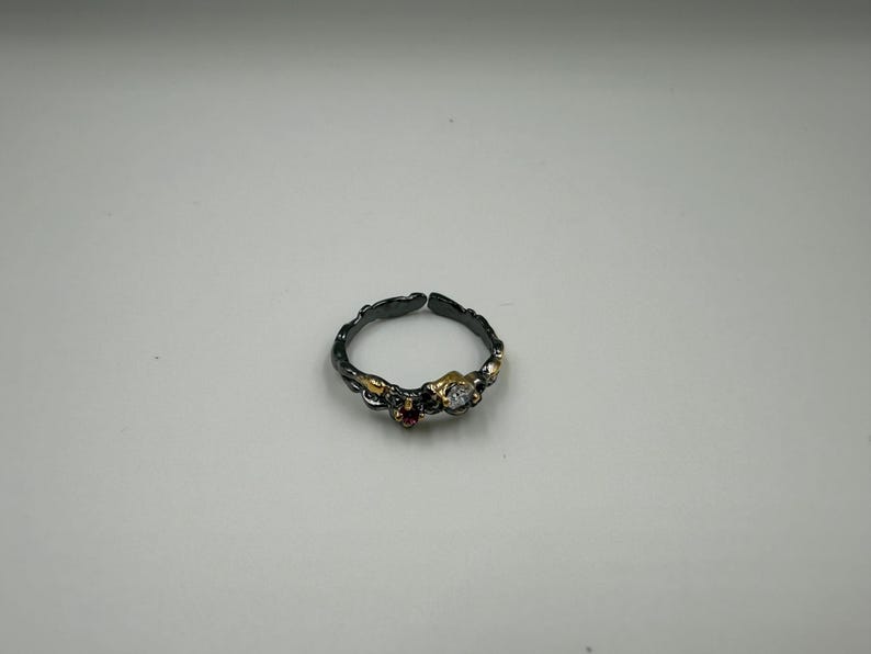 May include: An adjustable ring with a dark, textured band. The ring features gold accents and three gemstones: a clear, round stone and a small red stone. The ring is set against a white background.