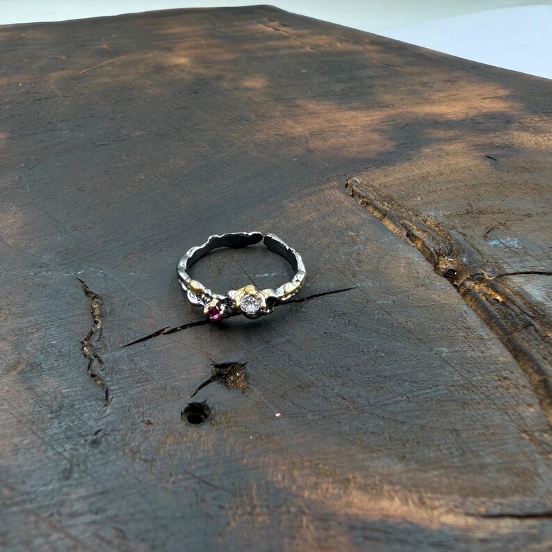 May include: A silver-toned ring with a textured band and decorative accents. The ring features a small, round, clear gemstone and a smaller, red gemstone. The ring is set on a dark, wooden surface.
