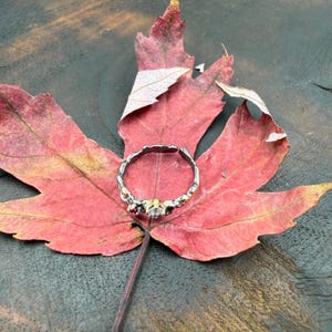 May include: A silver and gold ring with red and clear gemstones rests on a vibrant red maple leaf. The ring has a textured band and is open at the back. The leaf is set against a dark wood surface.