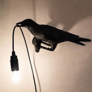Könnte beinhalten: Eine schwarze, rabenförmige Wandlampe mit einer Glühbirne. Der Rabe sitzt auf einem Ast und hält das Kabel der Lampe in seinem Schnabel. Die Lampe ist an einer weißen Wand befestigt, im Hintergrund sind ein Fenster und ein Vorhang zu sehen.