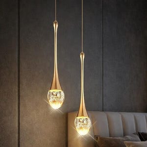 Może przedstawiać: Dwie złote lampy wiszące z kloszem w kształcie łzy. Lampy wiszą nad łóżkiem z zagłówkiem i poduszkami w neutralnych odcieniach. Pokój ma nowoczesną, elegancką estetykę.