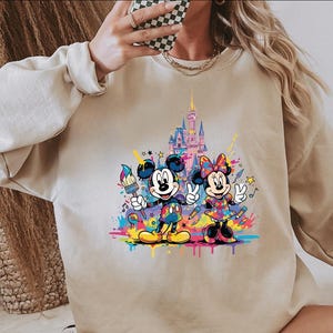May include: Beige sweatshirt featuring a colorful graphic of Mickey and Minnie Mouse with a castle backdrop. Mickey holds a paintbrush, Minnie makes a peace sign, and both are surrounded by paint splatters and stars.