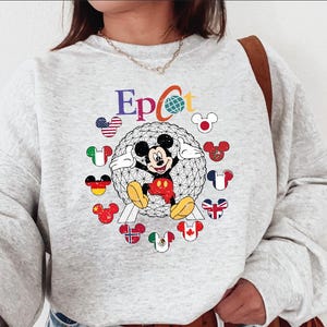 May include: A light gray sweatshirt featuring the word "Epcot" in colorful letters, with a globe graphic. Mickey Mouse is in the center, surrounded by Mickey Mouse heads with flags. The sweatshirt has long sleeves and a crew neck.