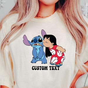 May include: Cream-colored t-shirt featuring a cartoon image of Stitch and Lilo. Lilo is kissing Stitch on the cheek. The text "CUSTOM TEXT" is printed below the image in a bold, black font. The shirt has short sleeves.